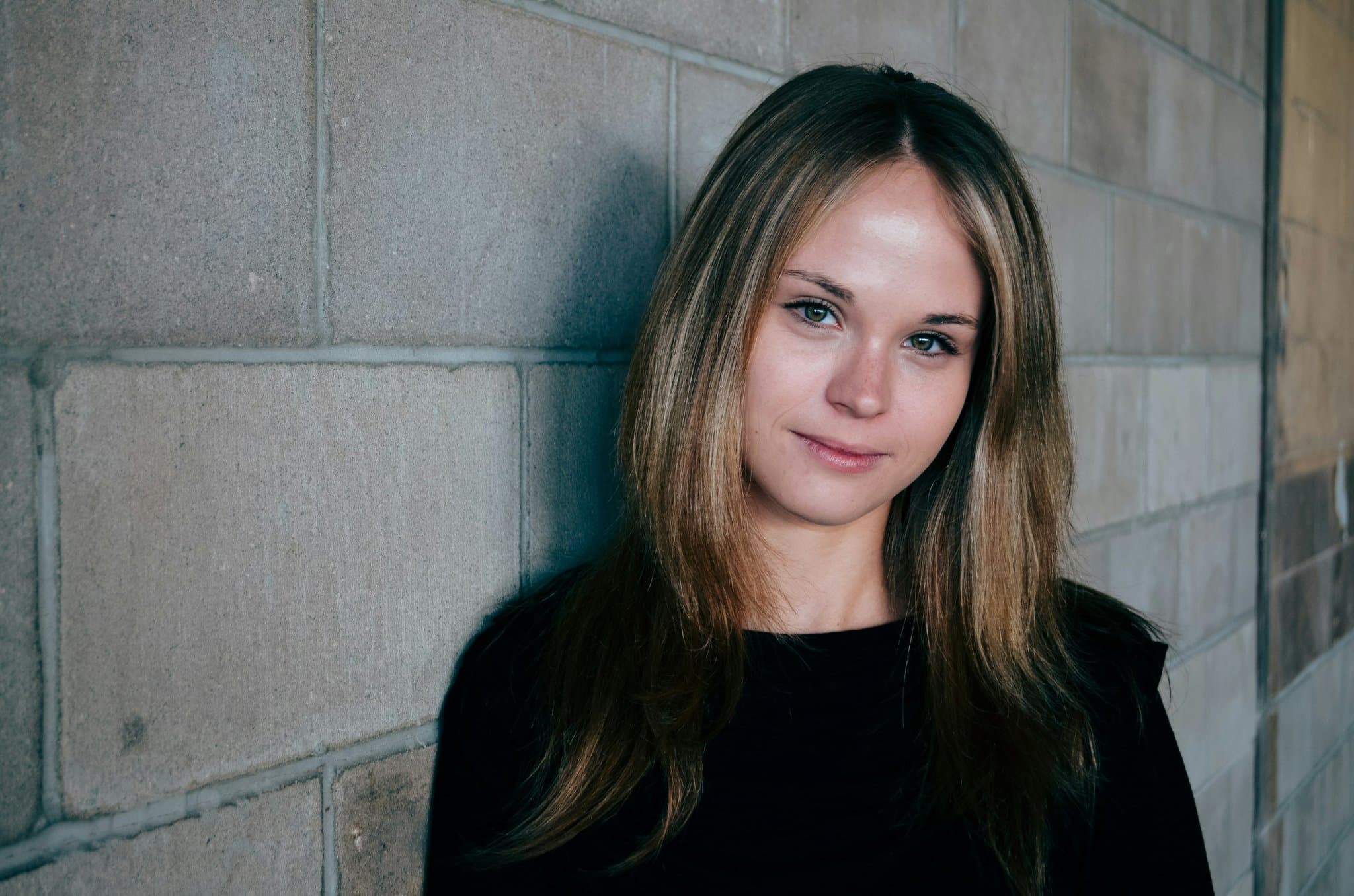 A woman leaning against a brick wall in simple casual wear.