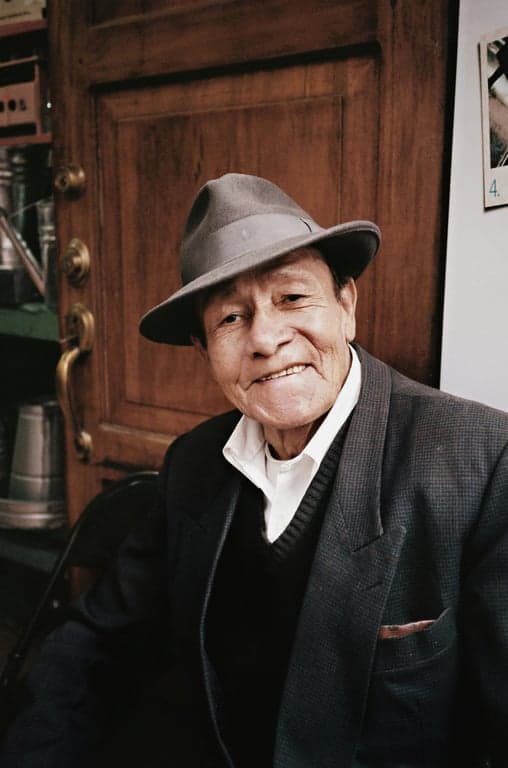 a portrait of a person wearing refined minimalist elderly menswear with a long tailored coat, relaxed straight silhouette, smooth wool or cotton fabric, understated construction, subtle layering, calm composed expression, soft directional lighting
