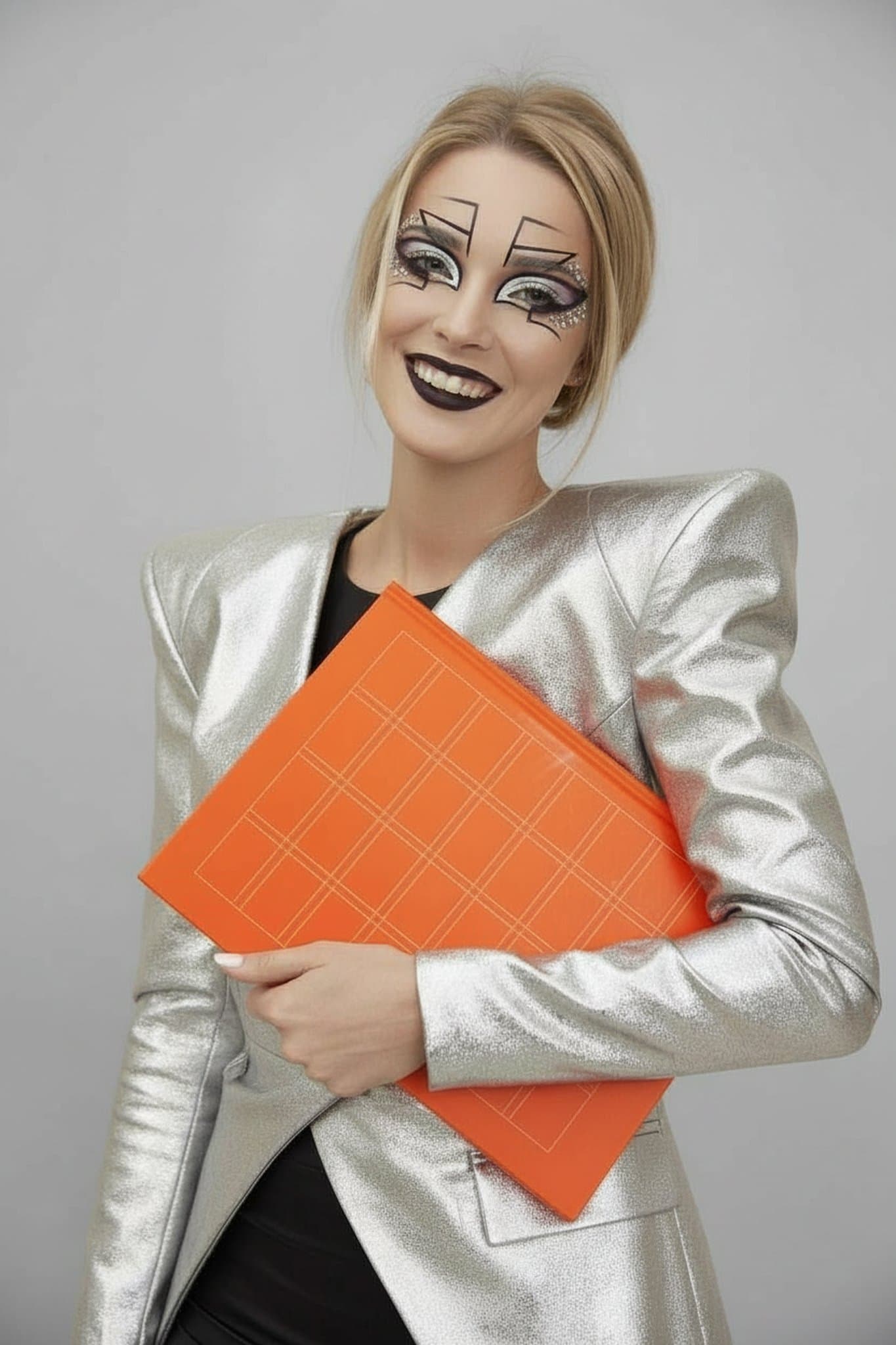 Silver structured jacket, bold geometric eye makeup, dark lipstick, holding bright orange folder.