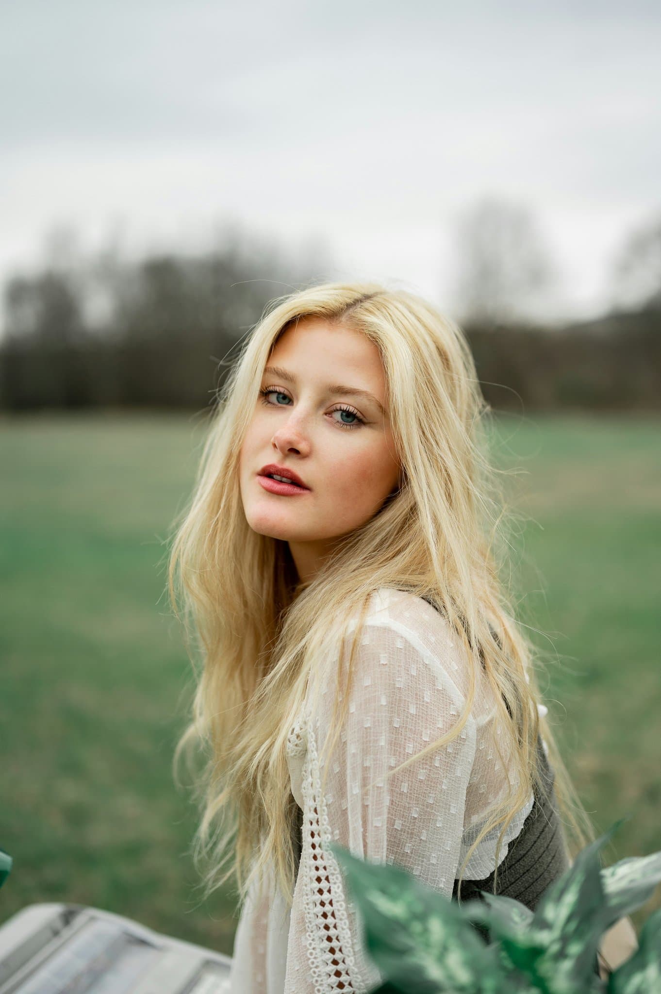 A woman outdoors with long blonde hair, wearing a light sheer blouse, posing against an open green field.