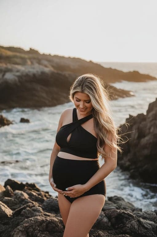 a portrait of a pregnant woman in a high-waisted maternity swimsuit, holding her belly by a rocky coastline