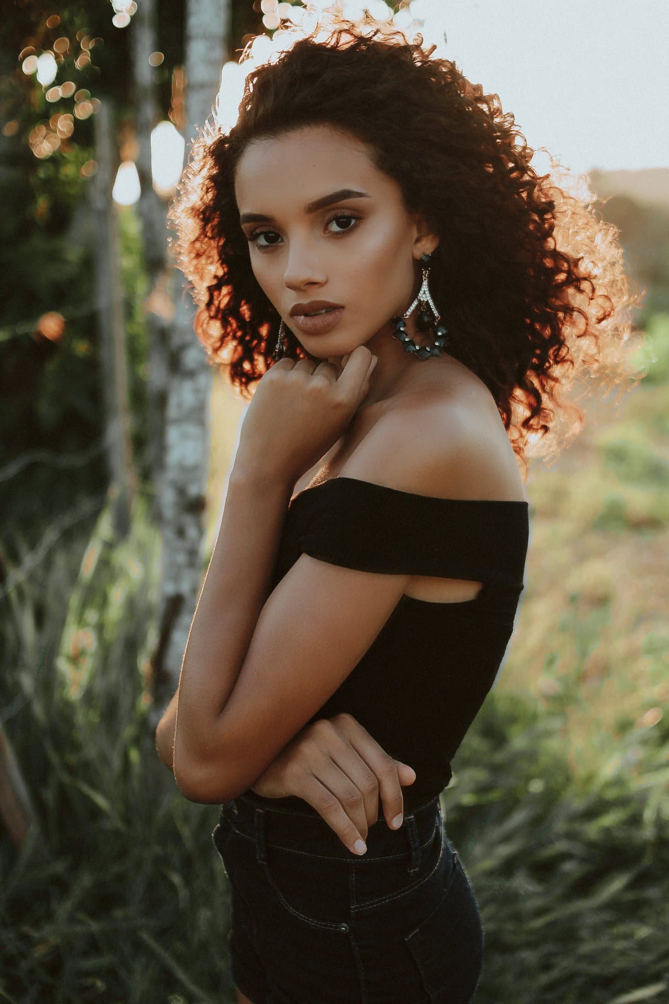 Woman in off shoulder black top posing outdoors at sunset with curly hair and soft golden light behind her.