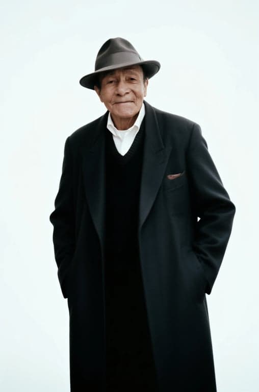 a portrait of a person wearing refined minimalist elderly menswear with a long tailored coat, relaxed straight silhouette, smooth wool or cotton fabric, understated construction, subtle layering, calm composed expression, soft directional lighting
