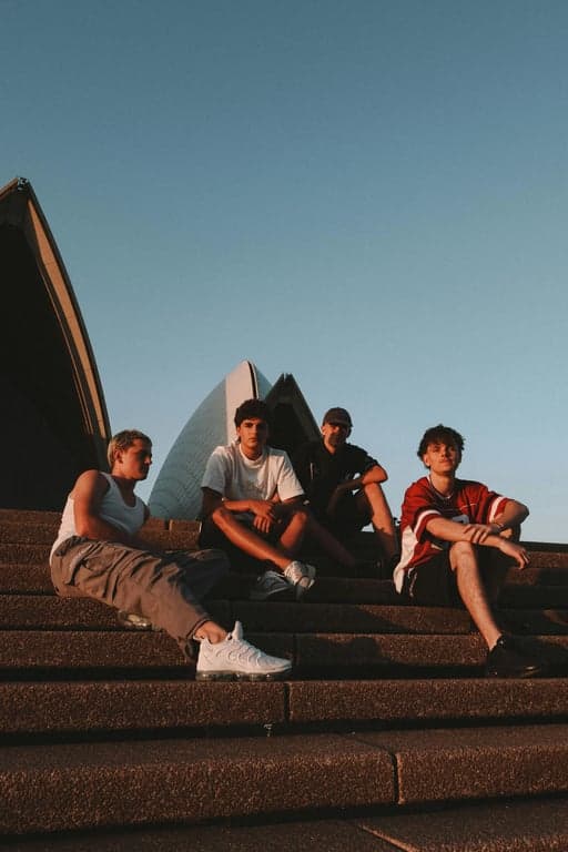 a portrait of multiple young men transformed into a modern K-pop boy group, wearing coordinated stage outfits with tailored jackets, fitted tops, slim trousers, intentional color harmony across members, clear outfit hierarchy, visible garment structure, clean fabric textures, confident upright posture, synchronized group presence, professional studio lighting, and a simple performance-style background