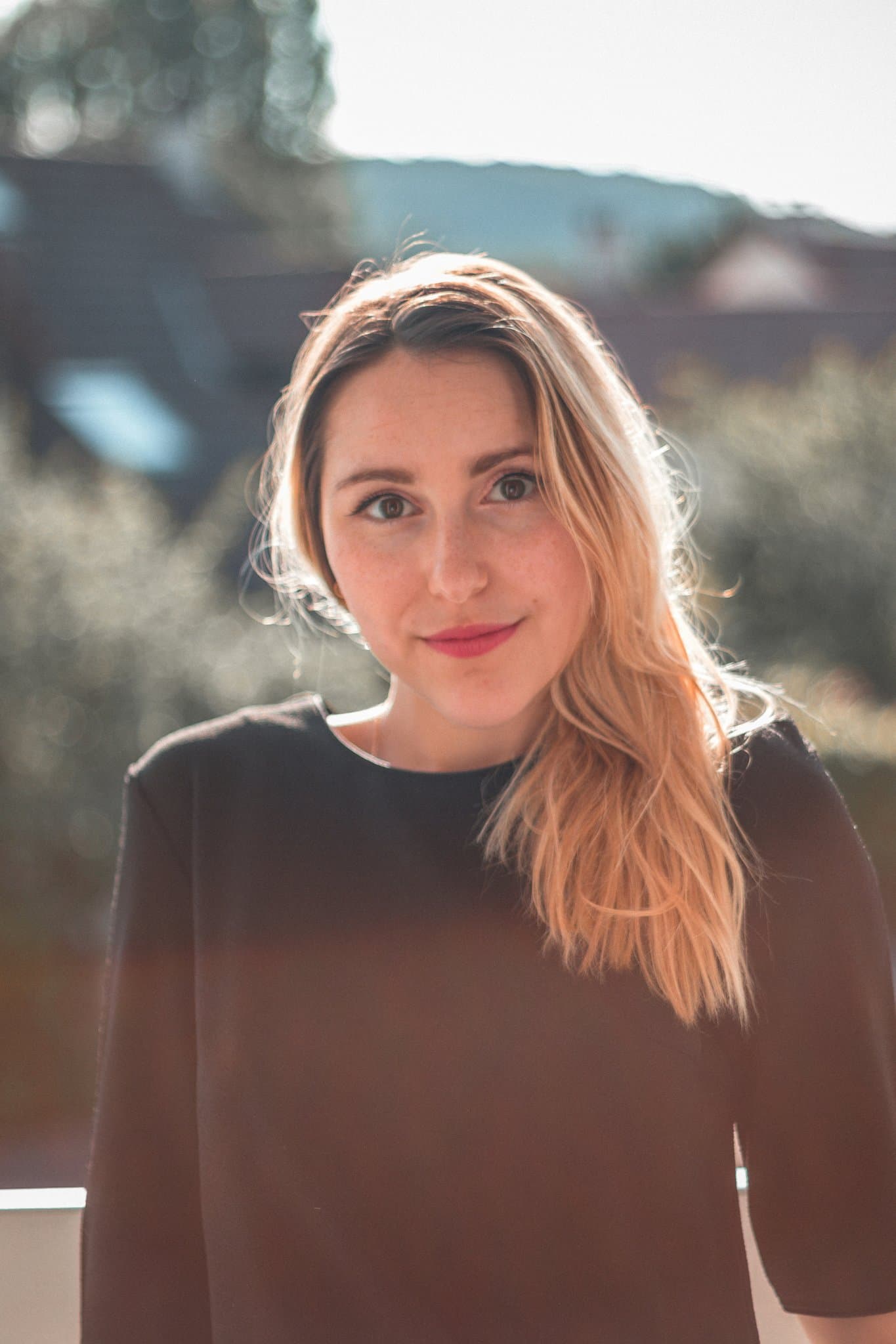 Outdoor portrait with soft sunlight, blonde hair, black top, gentle expression and warm background blur.
